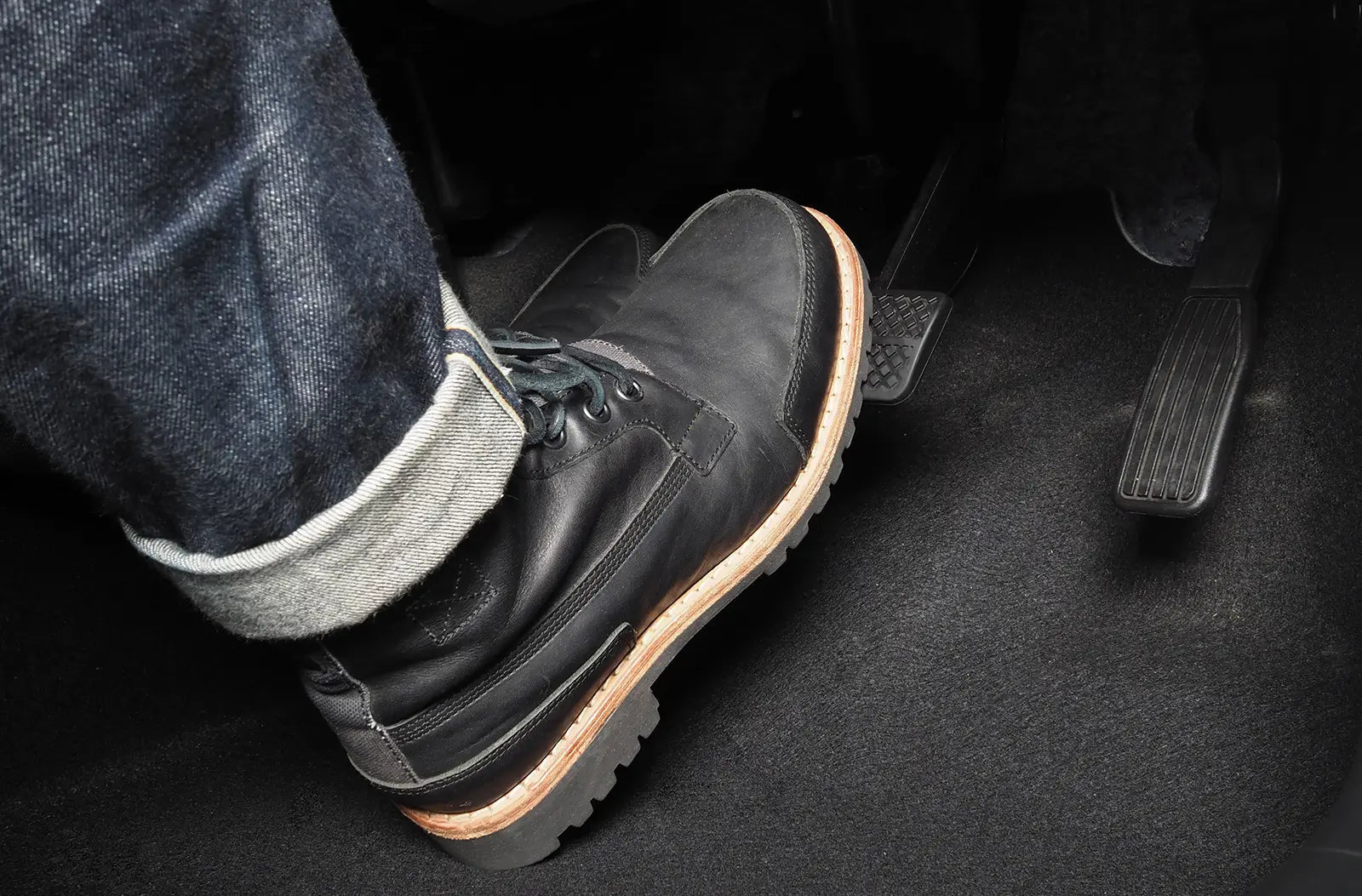 A close-up view from inside the footwell of a driver wearing a black leather boot pressing down hard on the vehicle's rubber brake pedal.