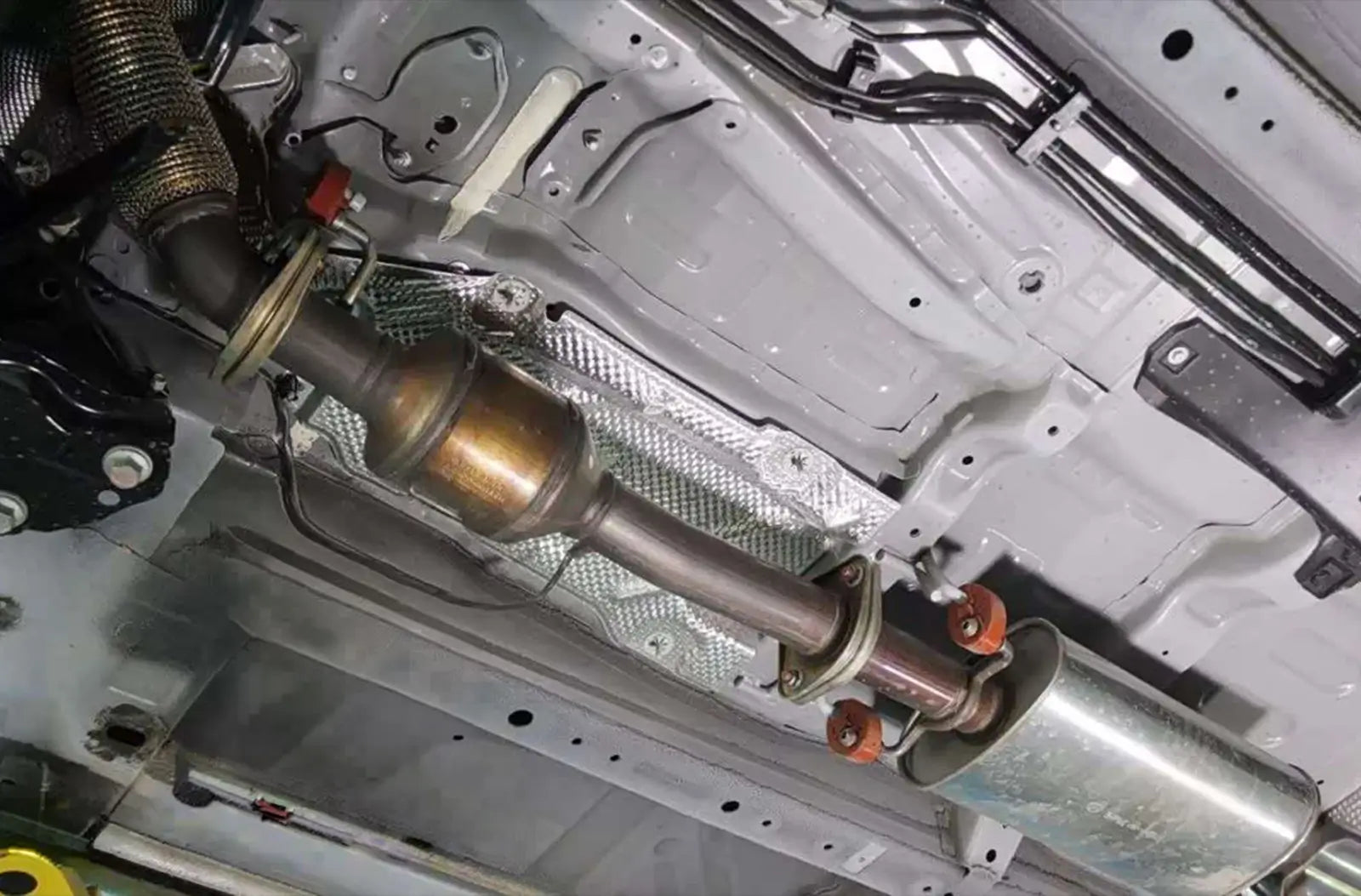 An under-car profile view of a vehicle's metallic exhaust pipe system, showcasing a threaded oxygen ($O_2$) sensor screwed directly into the catalytic converter piping.