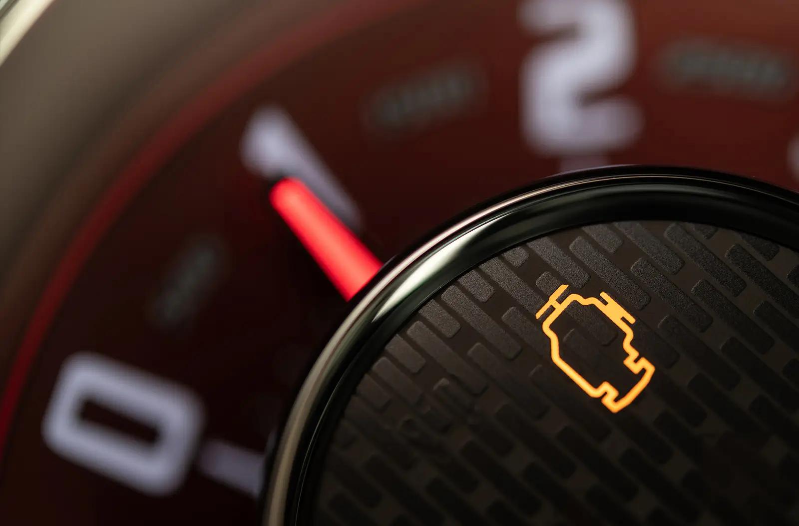 a close-up of a vehicle's dashboard, featuring an illuminated yellow check engine light symbol on a textured black circular element, with a blurred red tachometer needle and numbers in the background.
